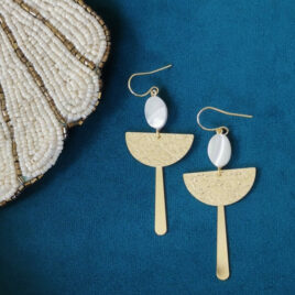 Boucles d'oreilles pendantes pour femme. dorées à l'or fin sur crochets classiques. en dessous desquels une pastille de nacre ovale et blanche pend, puis en dessous 2 éléments mis l'un sur l'autre : une forme demi lune martelée et une tige longue. modèle très graphique et épuré